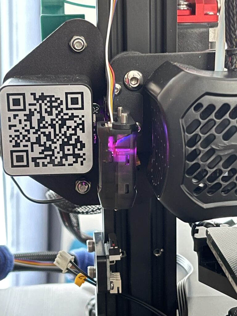 A detail of the print head of a Creality Ender 3 V2 3D-printer. In the middle of the picture once can see a square tube mounted at the print head. There is a cable going into the tube from the top and a purple light is glowing insight. At the bottom the top of the metail probe needle can be seen.