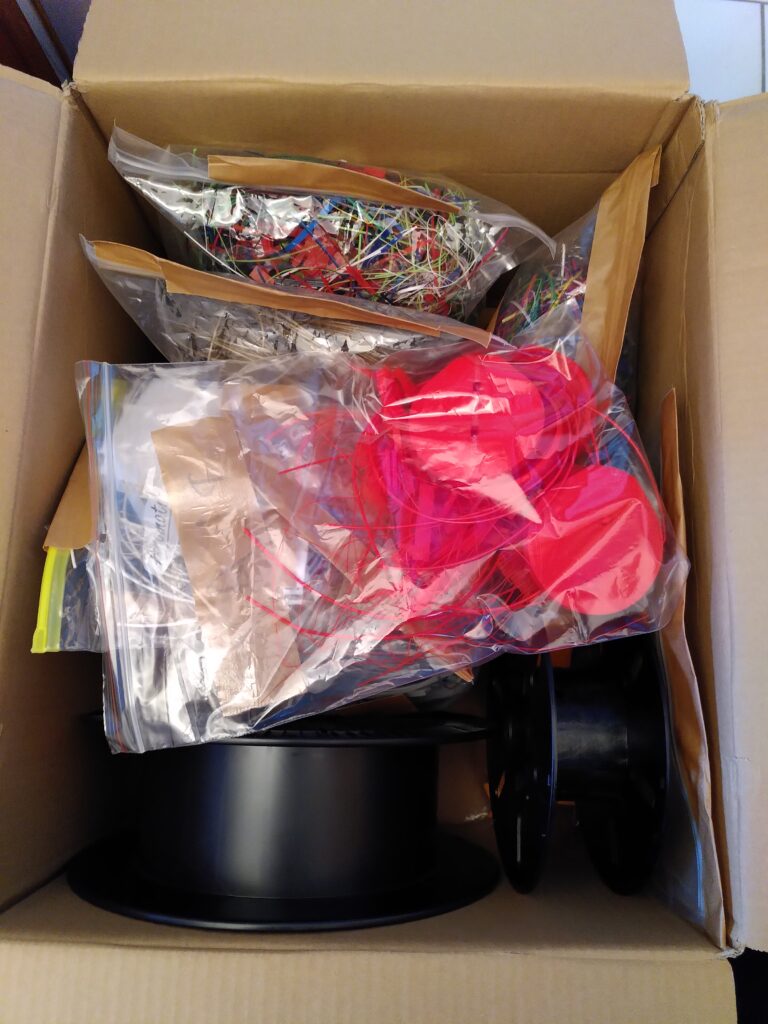 Ein großer Karton, darin einige leere Spulen und in Tüten verpackt, farblich sortierte Druckreste