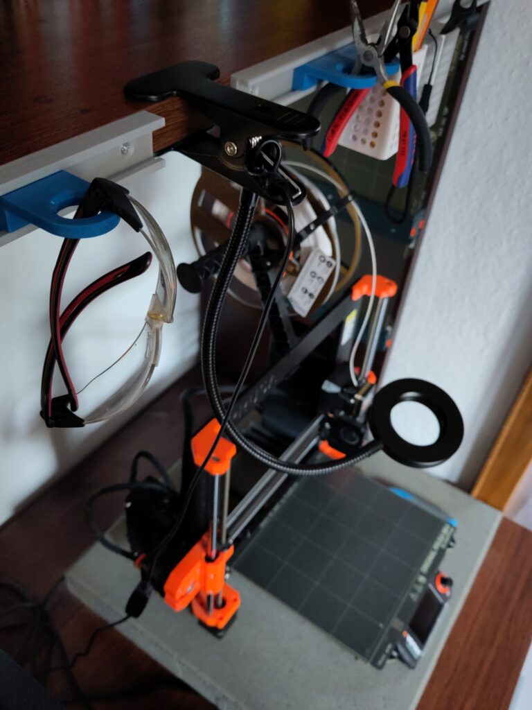 Tools hang on a toolbar above a 3D printer. There are also safety goggles hanging on it.